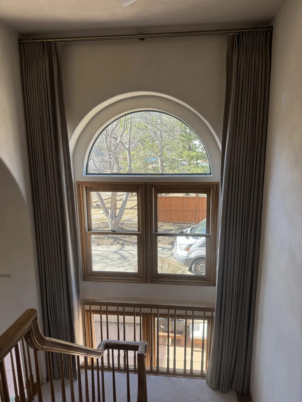 Custom gray pinch-pleat drapery panels on a brass rod framing an arched stairwell window, Howard's Draperies, Boulder